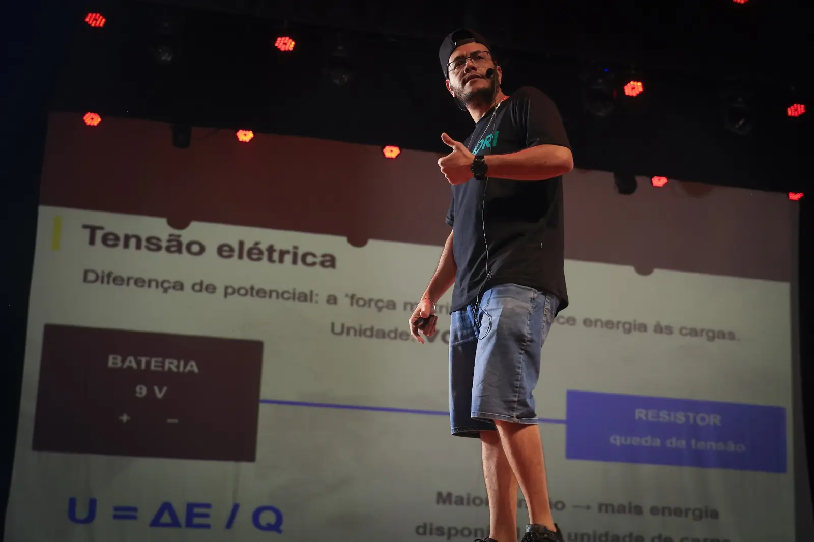 Professor da Ohmori explicando tensão elétrica no palco, com o conteúdo projetado no telão